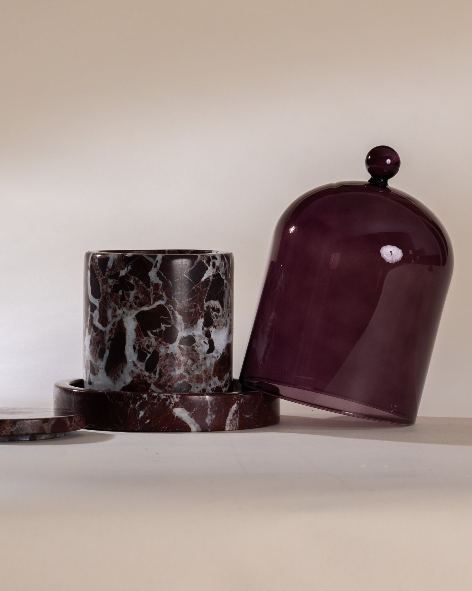 Marble-textured candle vessel and purple bell-shaped glass cloche on a neutral background