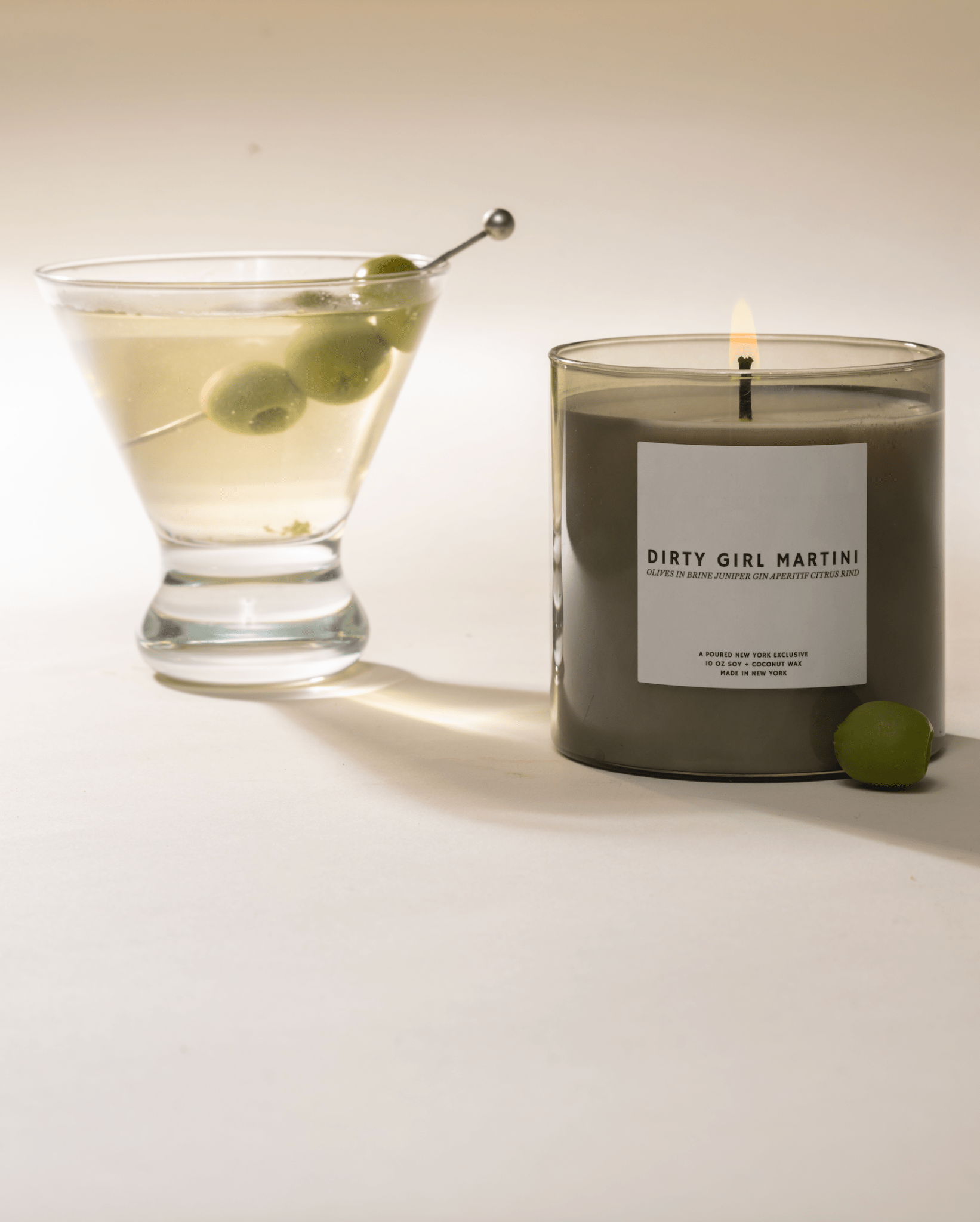Candle labeled 'Dirty Girl Martini' next to a glass of martini with olives on a white background