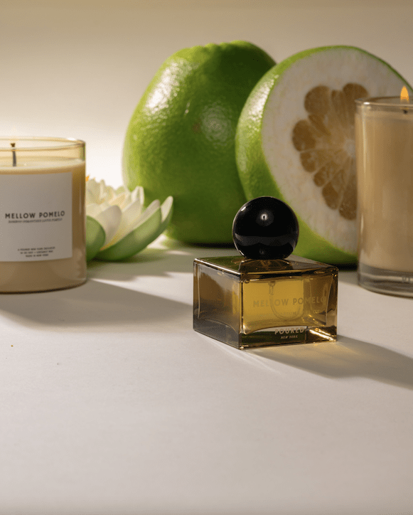 Poured New York 'Mellow Pomelo' lifestyle shot with the niche parfum in the foreground and the two candles in the background with a lotus flower and cut pomelo on neutral background.