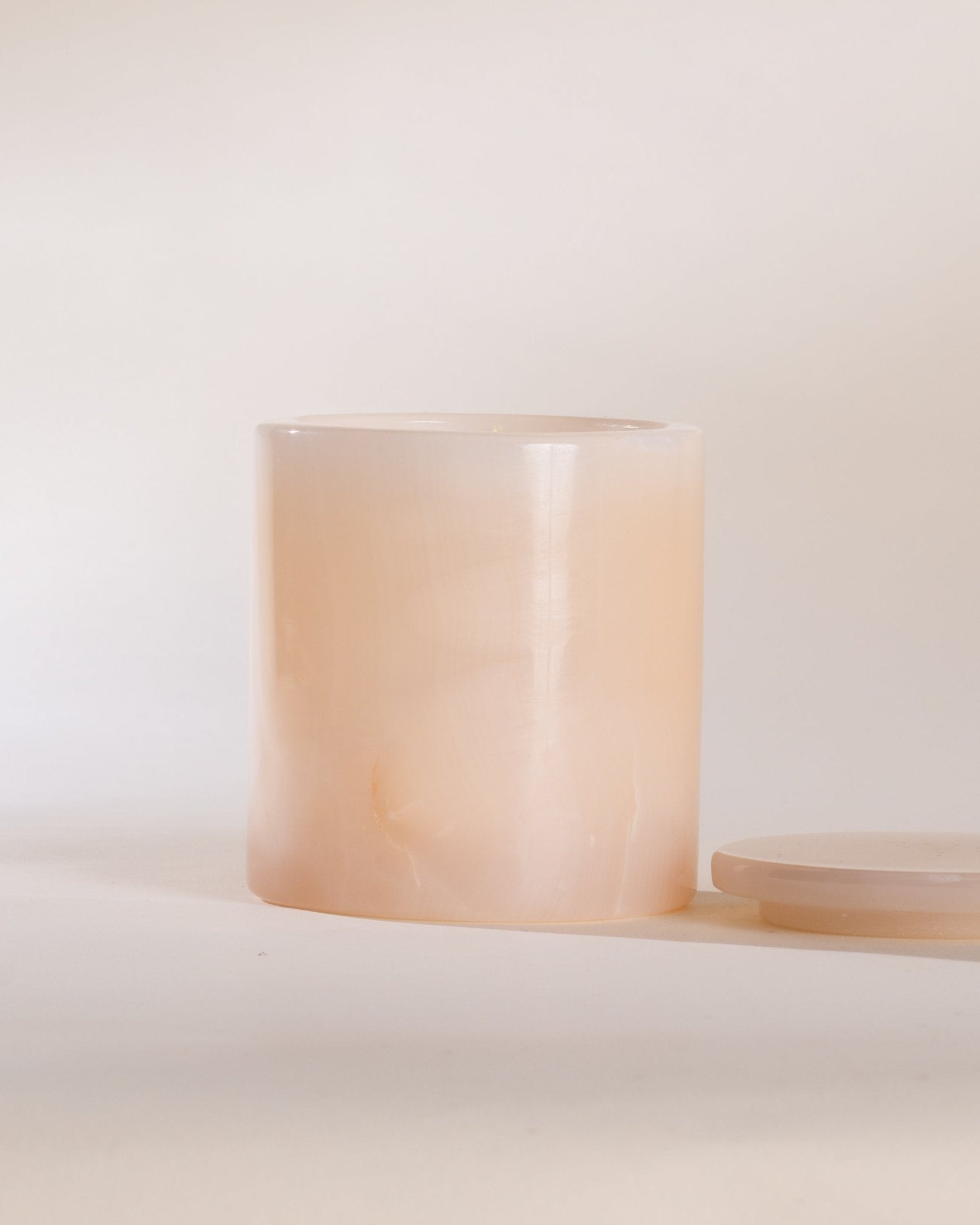 Cylindrical pink onyx candle vessel with a matching pink onyx lid on a white surface with a light beige background