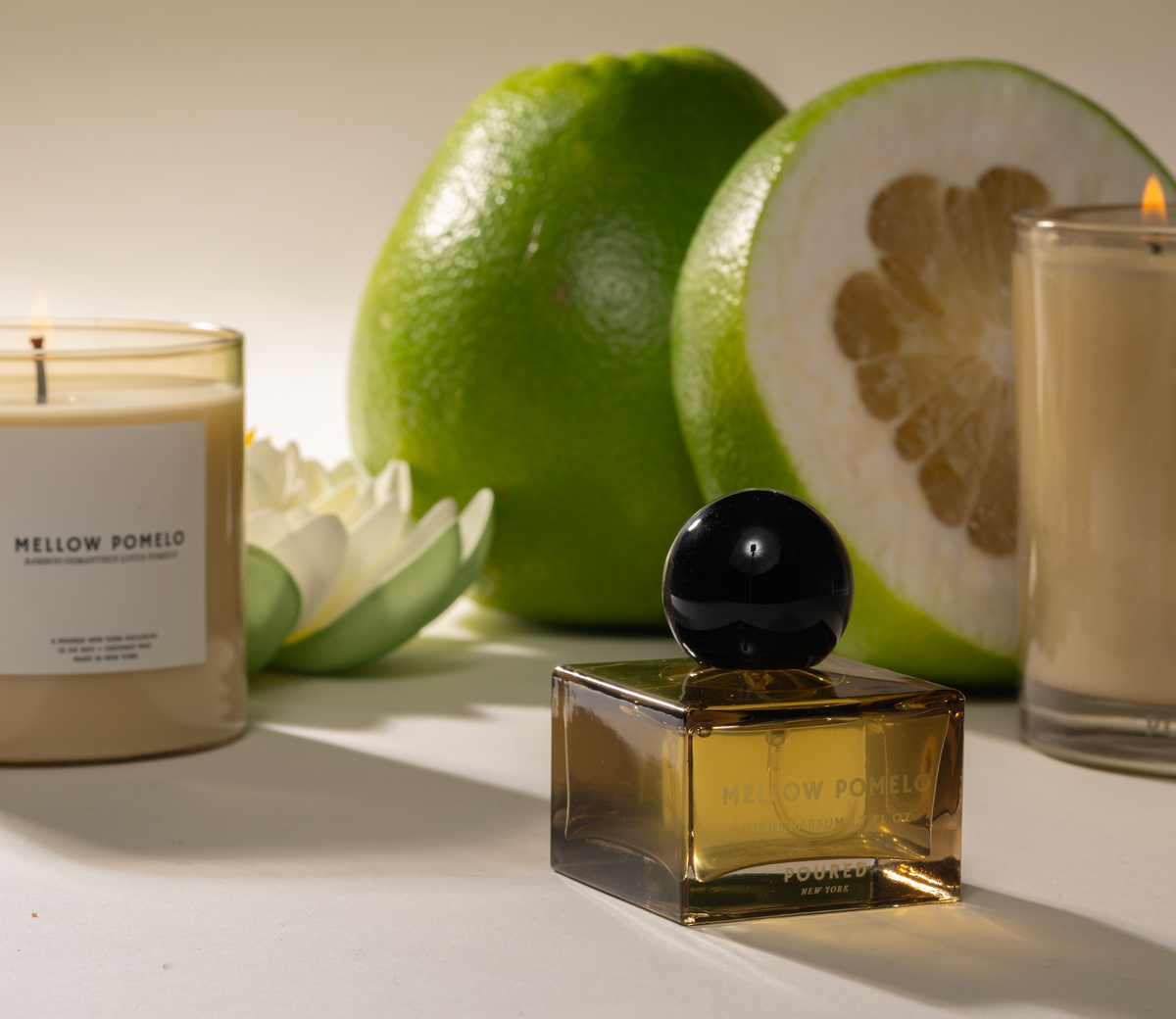 Mellow Pomelo square yellow perfume bottle, yellow single wick candle, and yellow double wick candle next to large green Pomelo fruit.