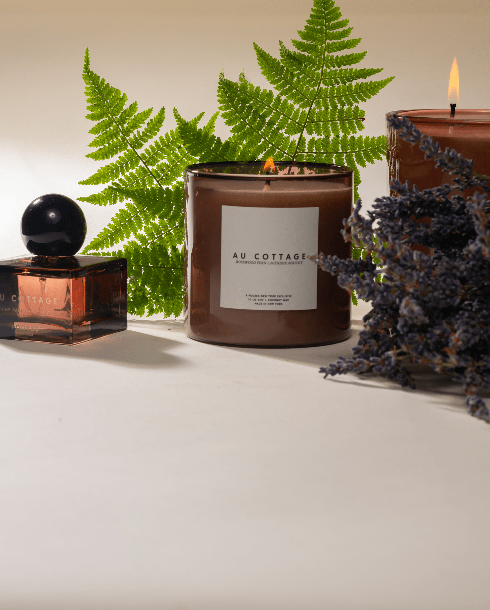 Au Cottage fragrances with two candles lit and a perfume with fern and lavender with a neutral background.
