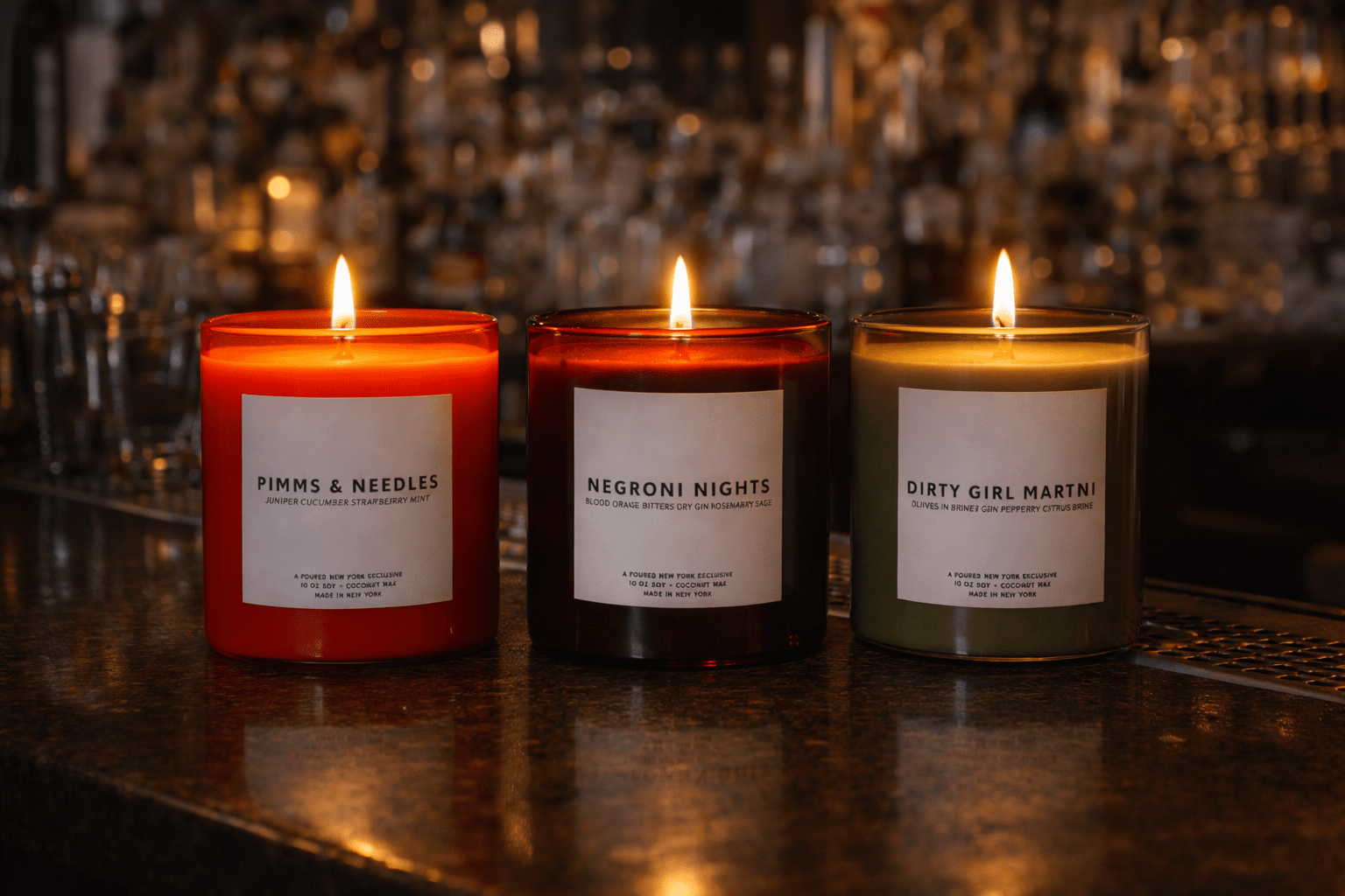 Three Poured New York candles lit on a bar featuring Pimms & Needles in a orange coral color, Negroni Nights in a dark red color, and Dirty Girl Martini in an olive color vessel. 