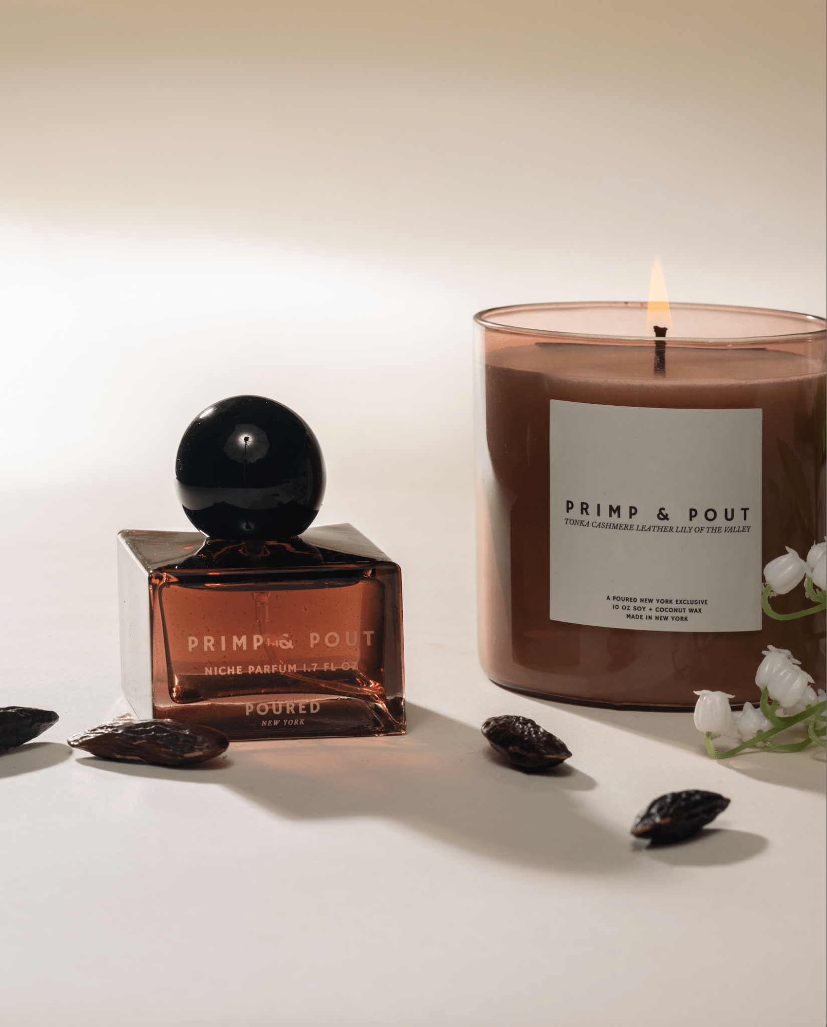Primp & Pout square perfume bottle and matching mauve candle with a white label and tonka beans and Lily of the Valley flowers on a neutral background.