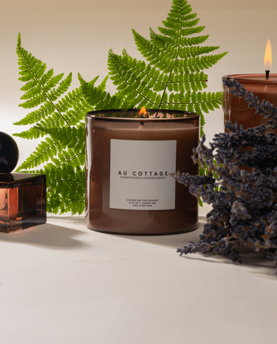 Two lit Poured New York 'Au Cottage' candles and a niche parfum with lavender and fern decor on a neutral background