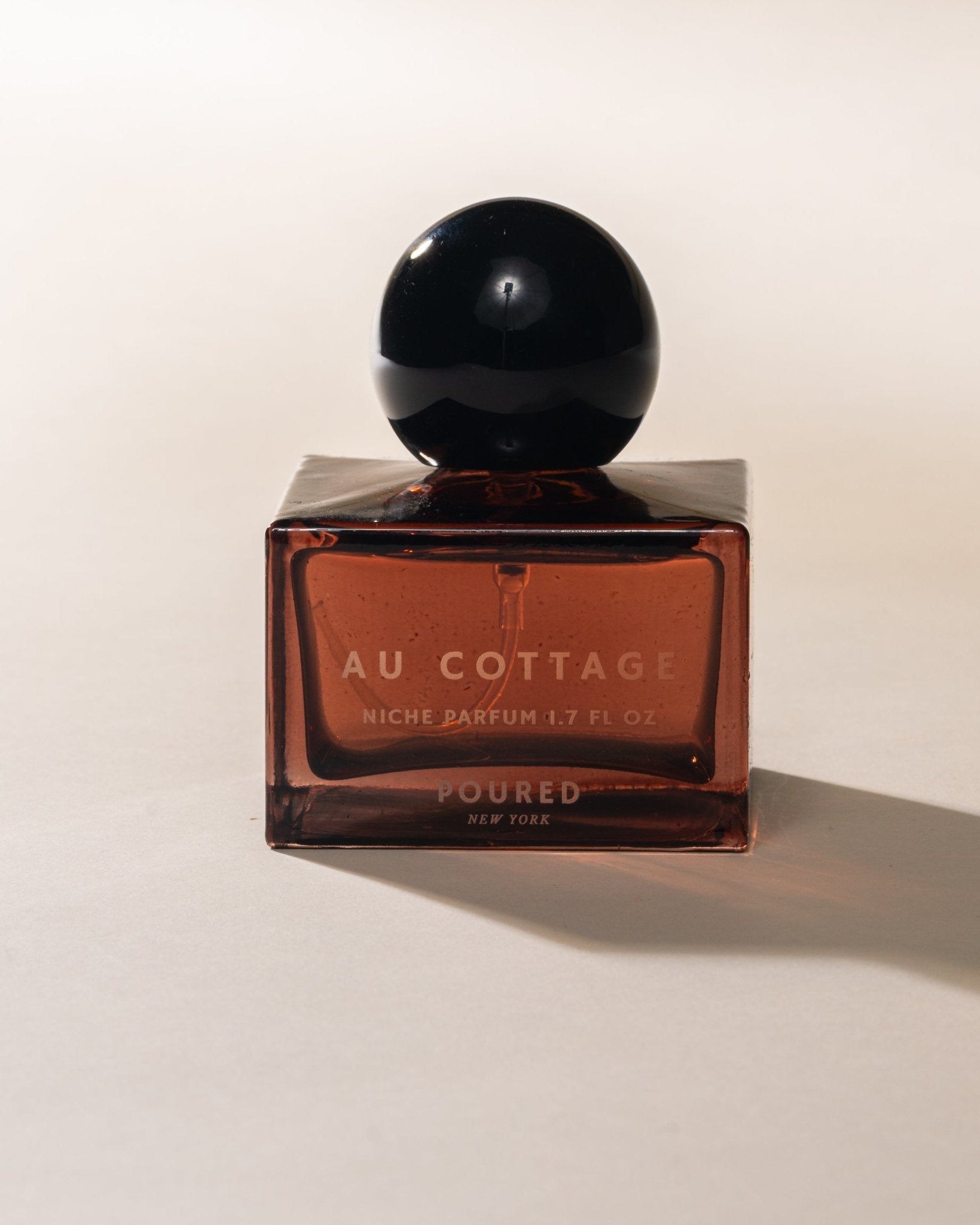 Perfume square bottle and black circular cap labeled 'Au Cottage' on a light background
