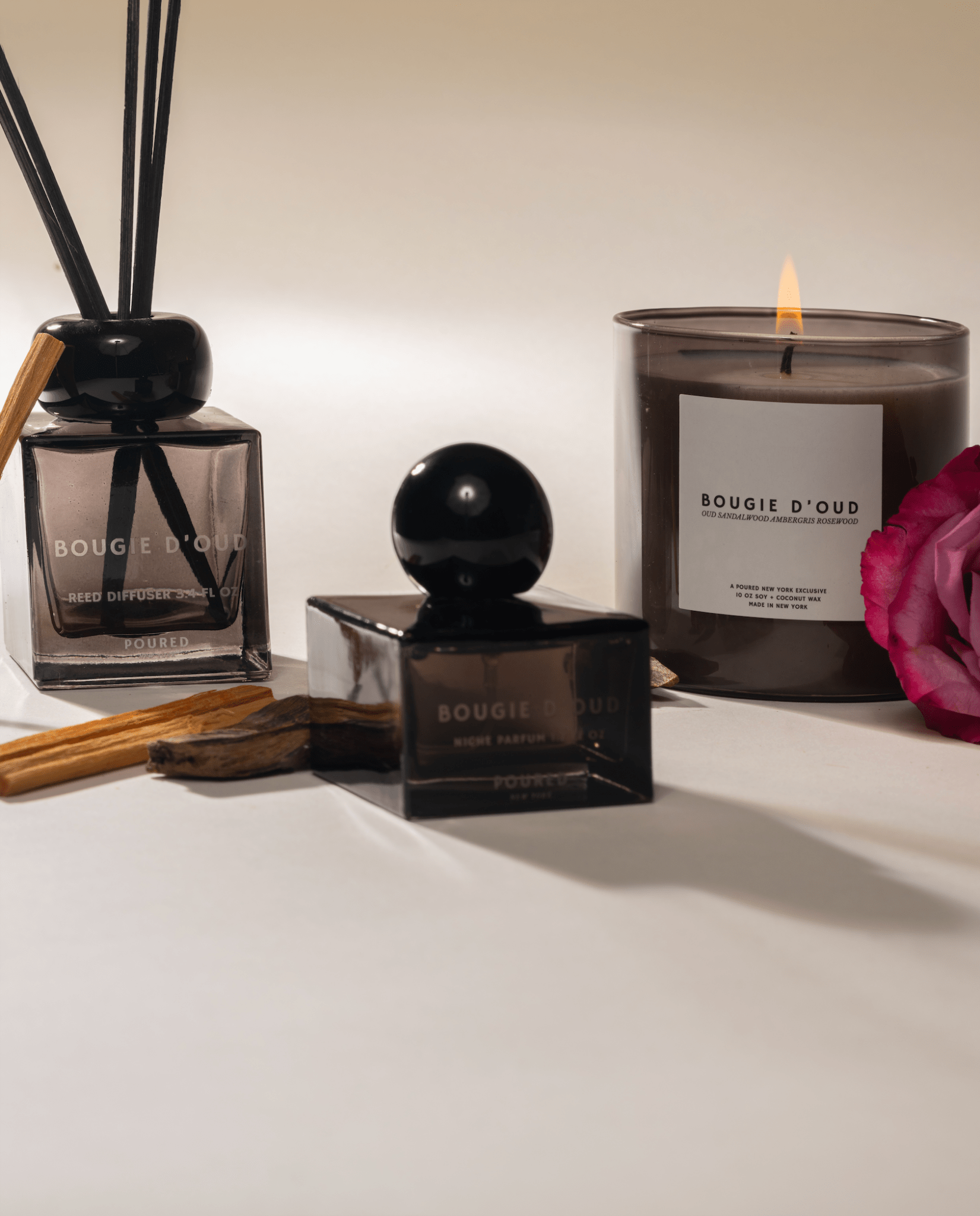 Bougie d'Oud candle, diffuser, and perfume bottle on a white surface with a pink rose, and sandalwood.