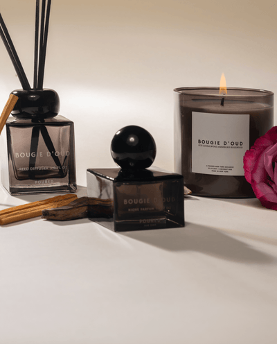Bougie d'Oud candle, diffuser, and perfume bottle on a white surface with a pink rose, and sandalwood.