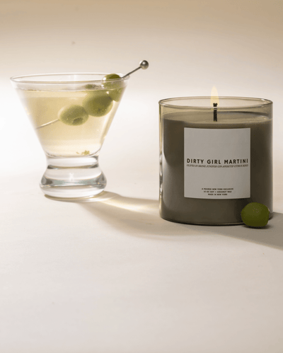 Candle labeled 'Dirty Girl Martini' next to a glass of martini with olives on a white background