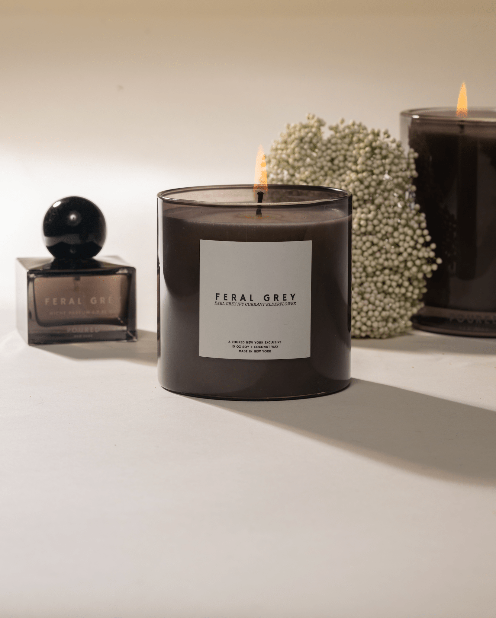 Poured New York 'Feral Grey' candles lit in grey glass and a grey niche parfum bottle with elderflower decor on a neutral background.