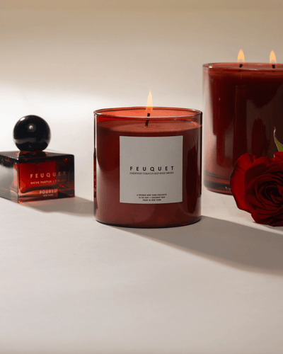 Two lit Feuquet candles and a perfume bottle on a light surface with a rose.