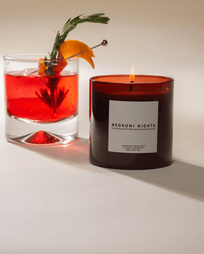Candle labeled 'Negroni Nights' next to a cocktail with an orange and rosemary garnish on a neutral background.