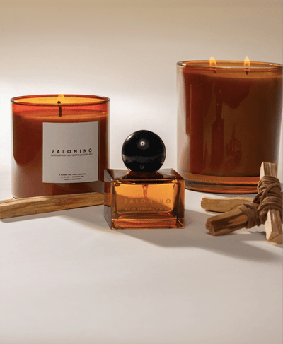 Poured New York 'Palomino' fragrances with a niche parfum glass square bottle in the foreground and the two candles in the background with palo santo and leather decor on a neutral background