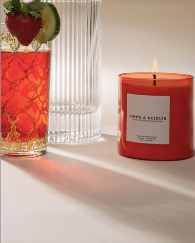 Red candle labeled 'PIMMS & NEEDLES' next to a glass with a strawberry and lime garnish on a light surface.