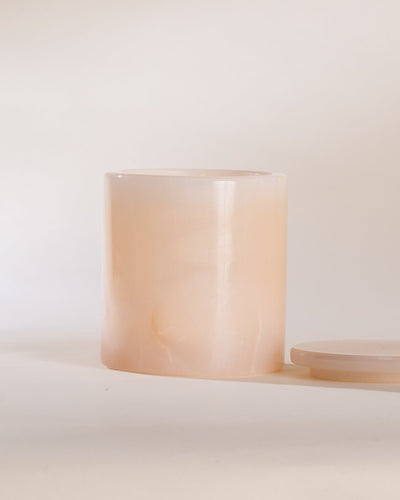 Cylindrical pink onyx candle vessel with a matching pink onyx lid on a white surface with a light beige background