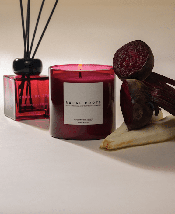 Red candle labeled 'Rural Roots' with a matching square diffuser and cut beets and white wild carrots on a neutral background