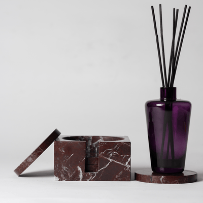 Dark Red Marble Square Coaster Set that comes with four matching dark red marble coasters on a neutral background with a purple hand blown glass reed diffuser sitting on the coaster. 